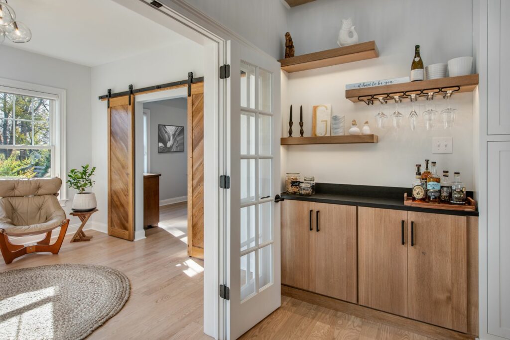 A wooden bar cabinet stands against a light wall, featuring a countertop with bottles and glasses. Soft light illuminates the space, revealing an adjacent room through glass-paneled doors.