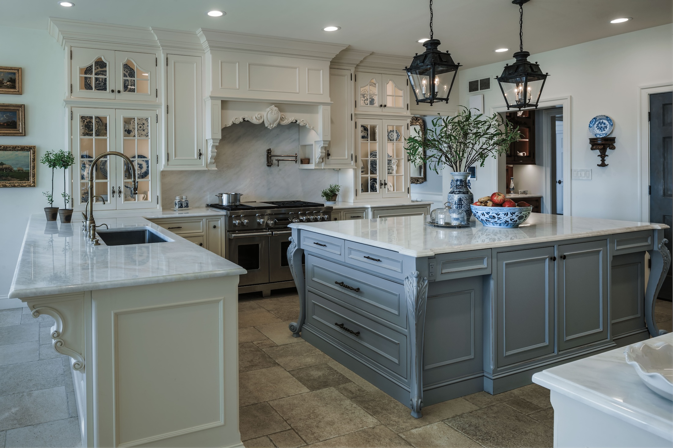 A spacious kitchen features a large marble island with drawers and a sink, a stainless steel stove, elegant cabinetry with glass doors, and decorative hanging lanterns, all in a bright, modern design.