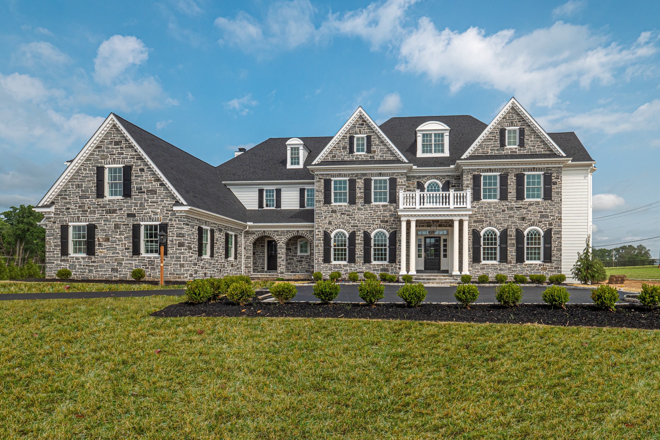 A large, elegant house with stone and siding features stands prominently. Its porch has white columns, and the landscaped front yard includes neatly trimmed bushes, set against a blue sky.