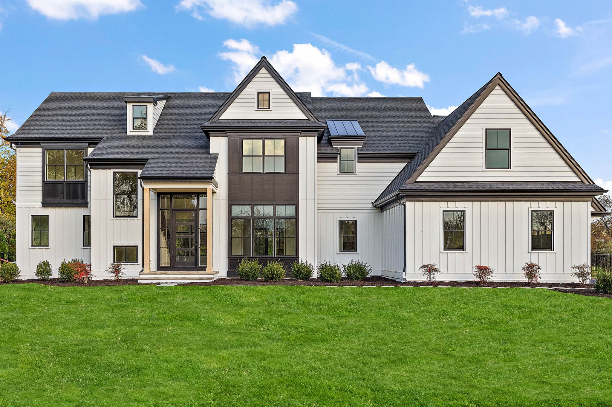 A modern, two-story house features a mix of white and dark siding, large windows, and a prominent front porch, surrounded by a green lawn and landscaped shrubs under a blue sky.