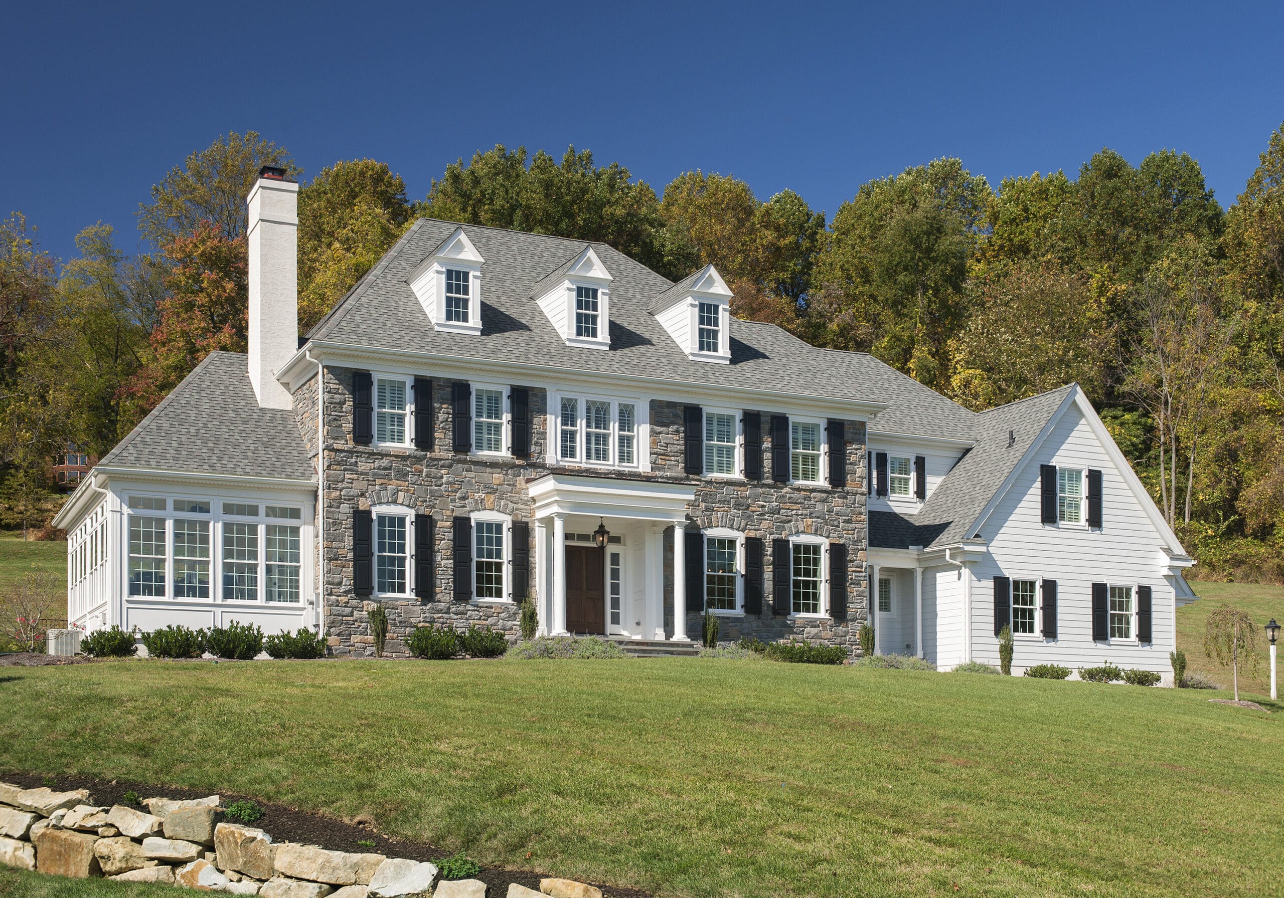 A two-story, colonial-style house features a stone facade, white clapboard siding, and black shutters. It is set on a green lawn surrounded by trees under a clear blue sky.