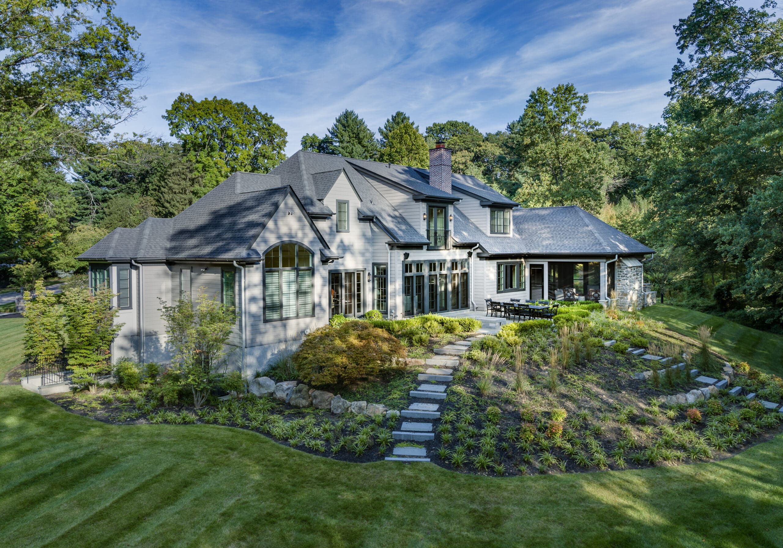 A large, modern house with a sloped roof is set on a landscaped yard featuring stone pathways, shrubs, and trees, surrounded by greenery under a clear blue sky.