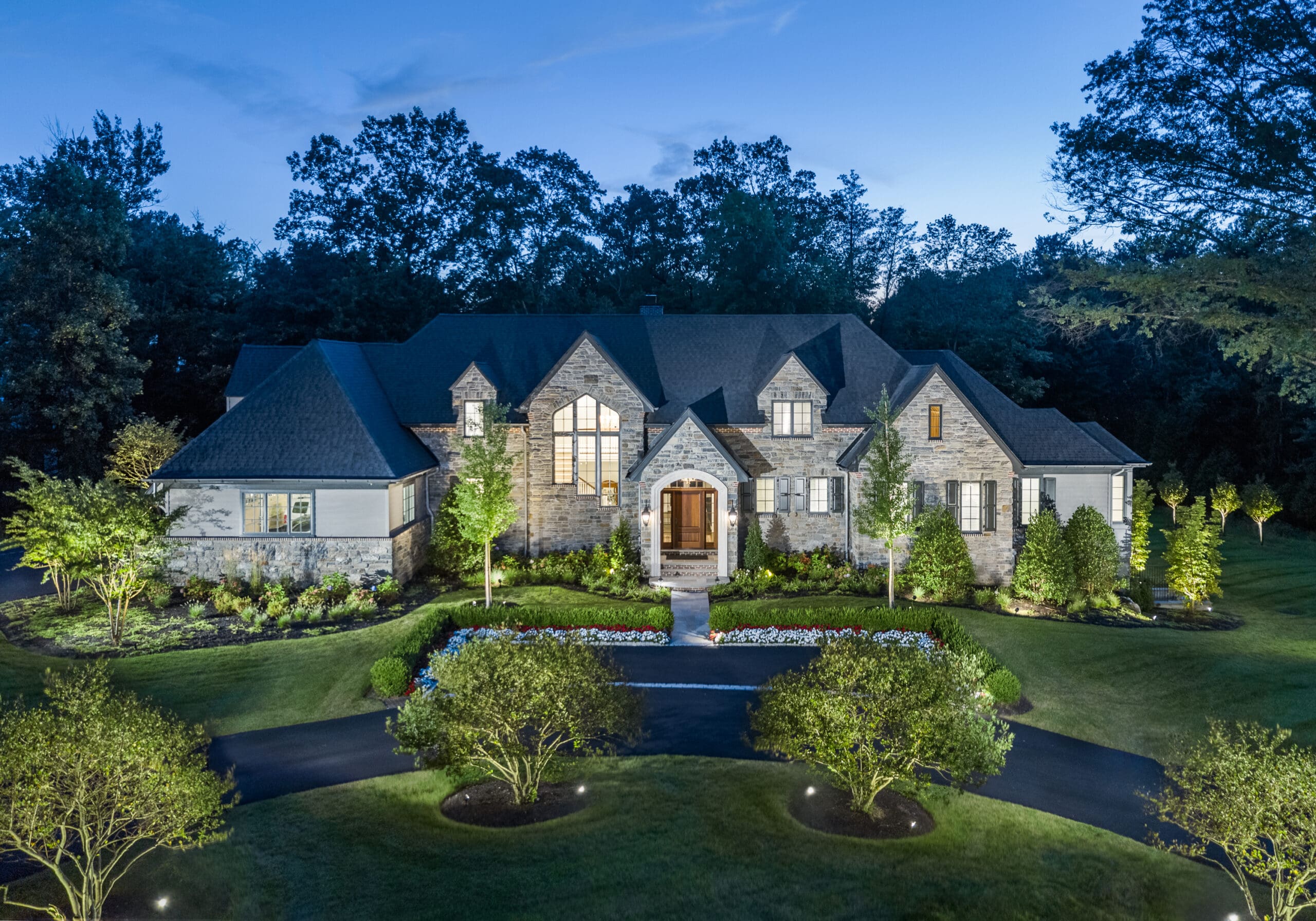 A large, elegantly designed stone house stands illuminated at dusk. Surrounding it are well-manicured gardens and trees, with a winding driveway leading to the entrance.