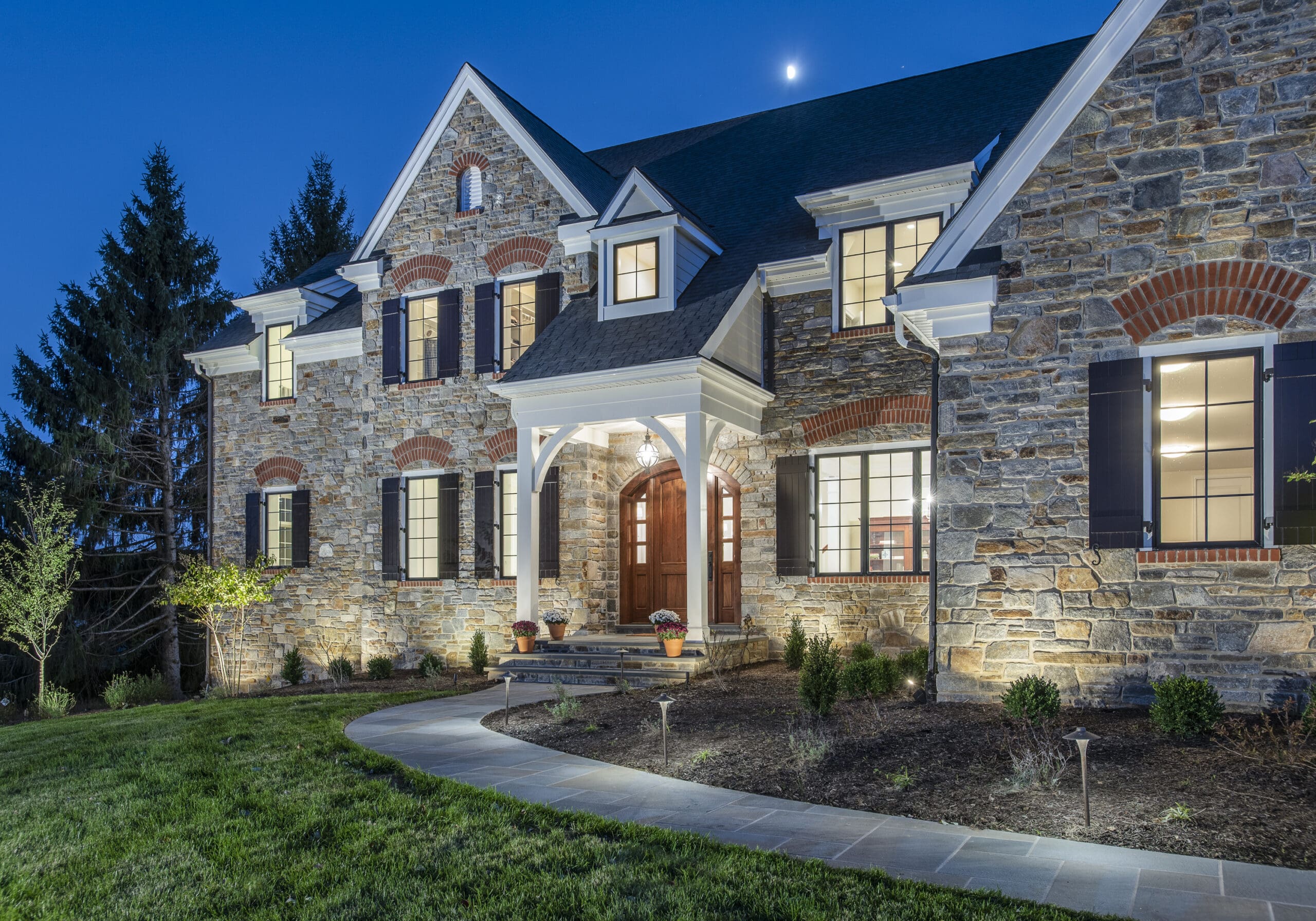 A large stone house is illuminated at dusk, featuring a welcoming entrance with steps leading to a wooden door, surrounded by landscaped greenery and soft outdoor lighting.