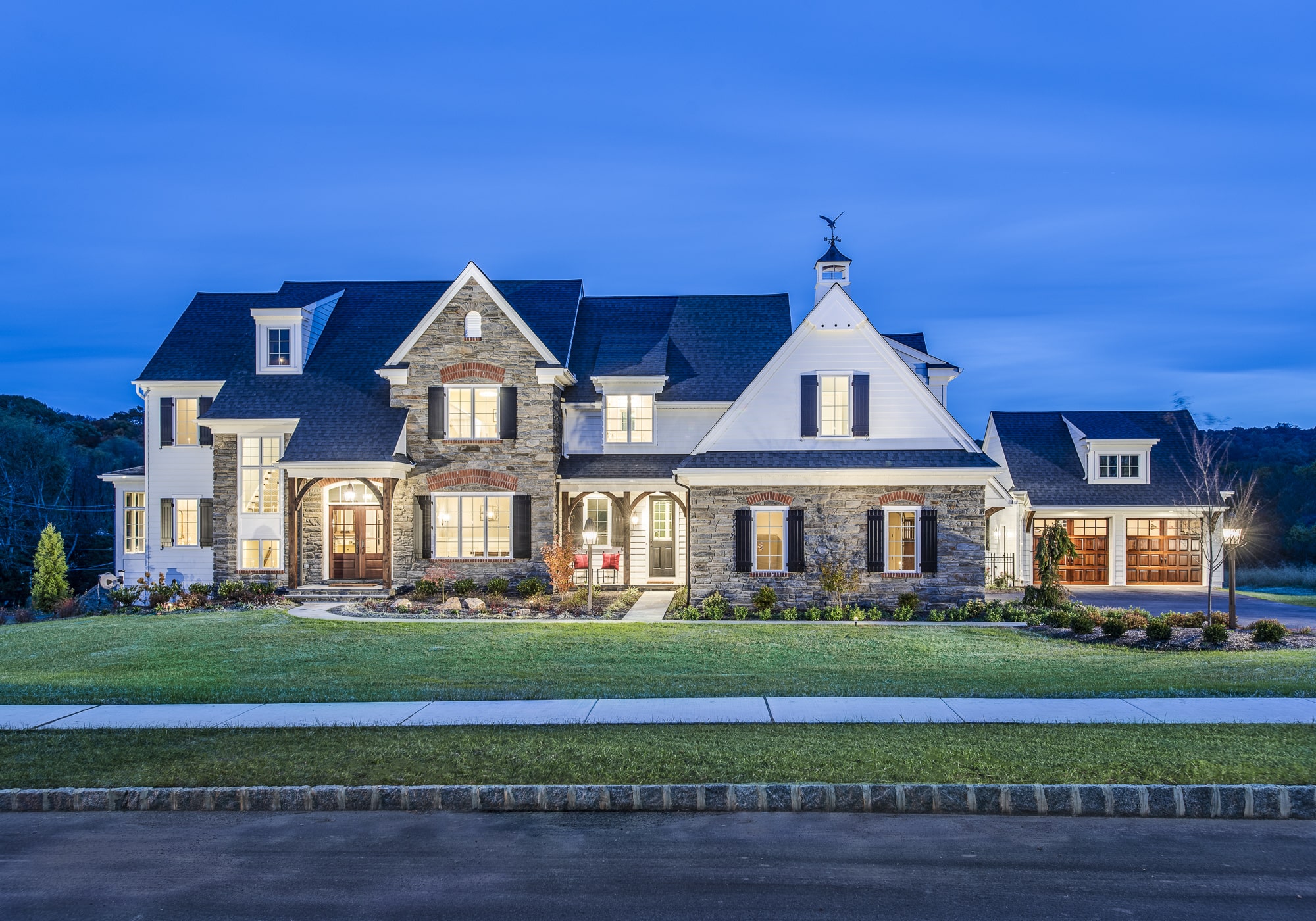 A large, modern house with stone and siding features stands prominently, illuminated at dusk. Surrounding greenery and a paved walkway enhance its inviting appearance in a serene residential area.