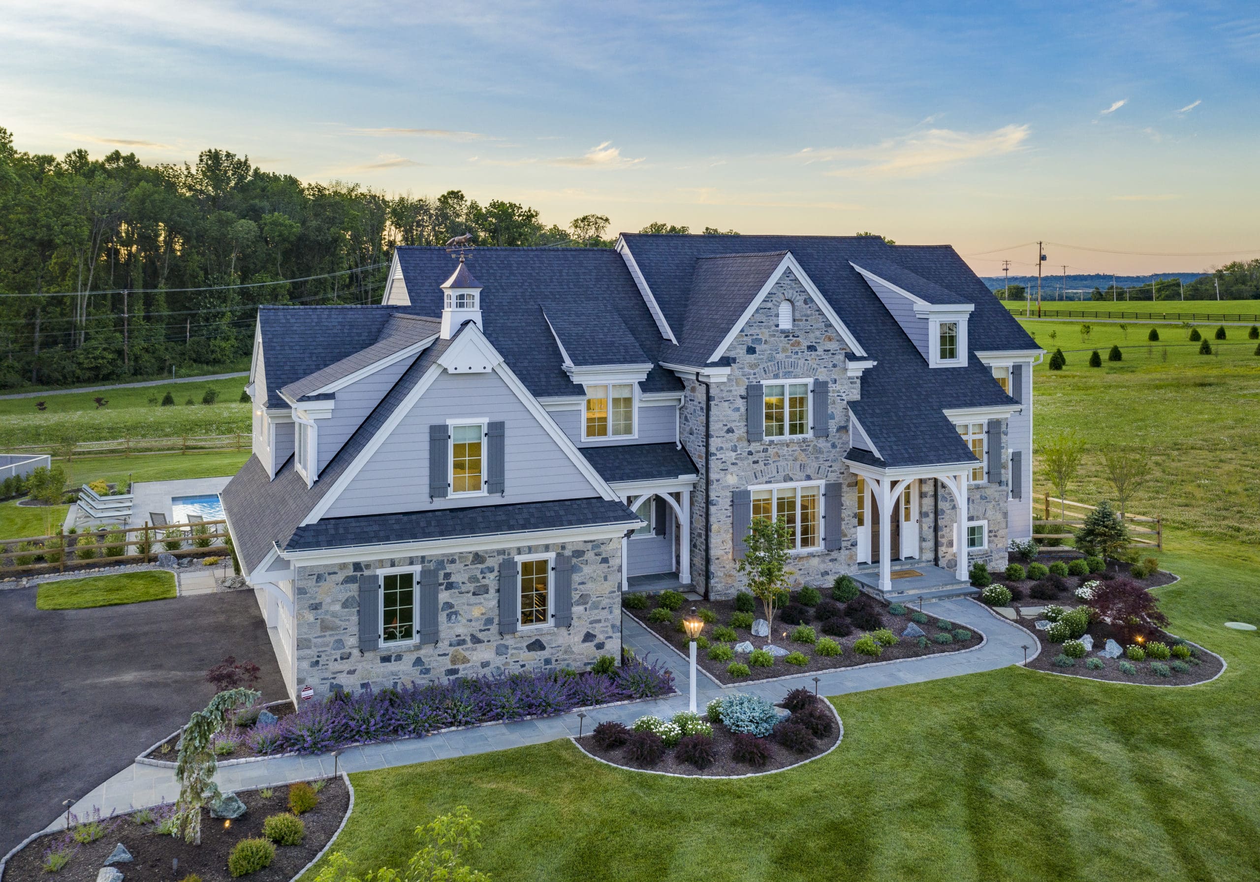 A large, modern home with stone and siding features stands amid a lush green lawn. The house is surrounded by landscaped gardens, with a pool visible in the background.