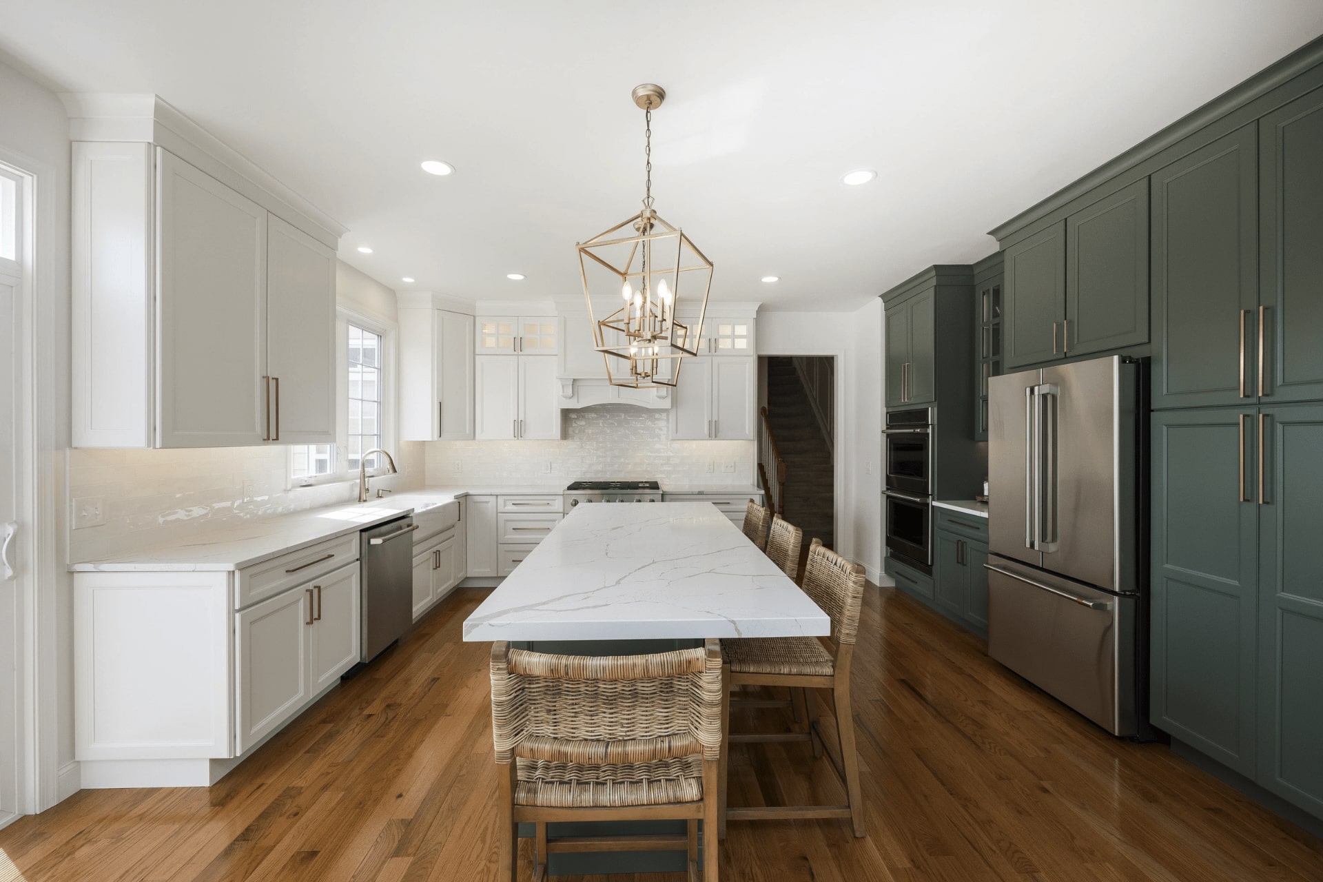 A modern kitchen features white and dark green cabinetry, a large marble island with woven bar stools, stainless steel appliances, and a stylish overhead light, all set on wooden flooring.