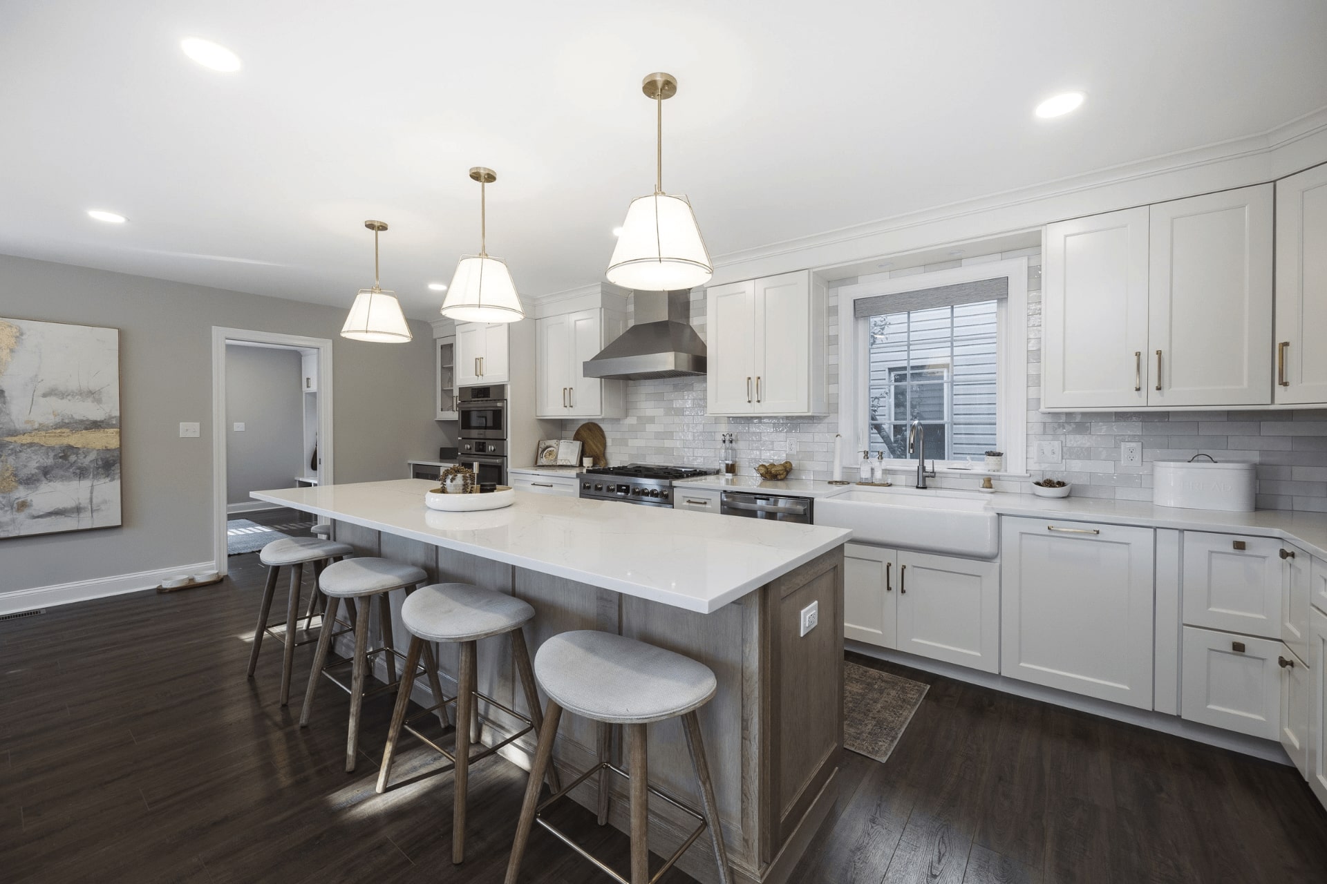 A modern kitchen features a central island with four stools. Pendant lights illuminate the white cabinetry and stainless steel appliances. Natural light enters through a window, enhancing the bright atmosphere.