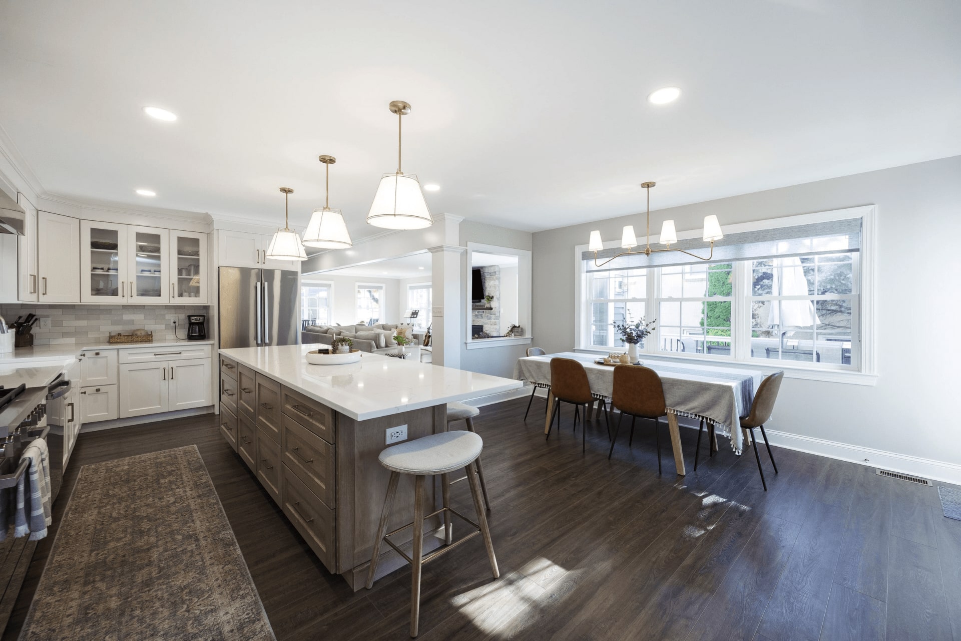 A modern kitchen features an island with a white countertop, bar stool, and wooden cabinets. Elegant lighting hangs above, with a dining area nearby and large windows letting in natural light.