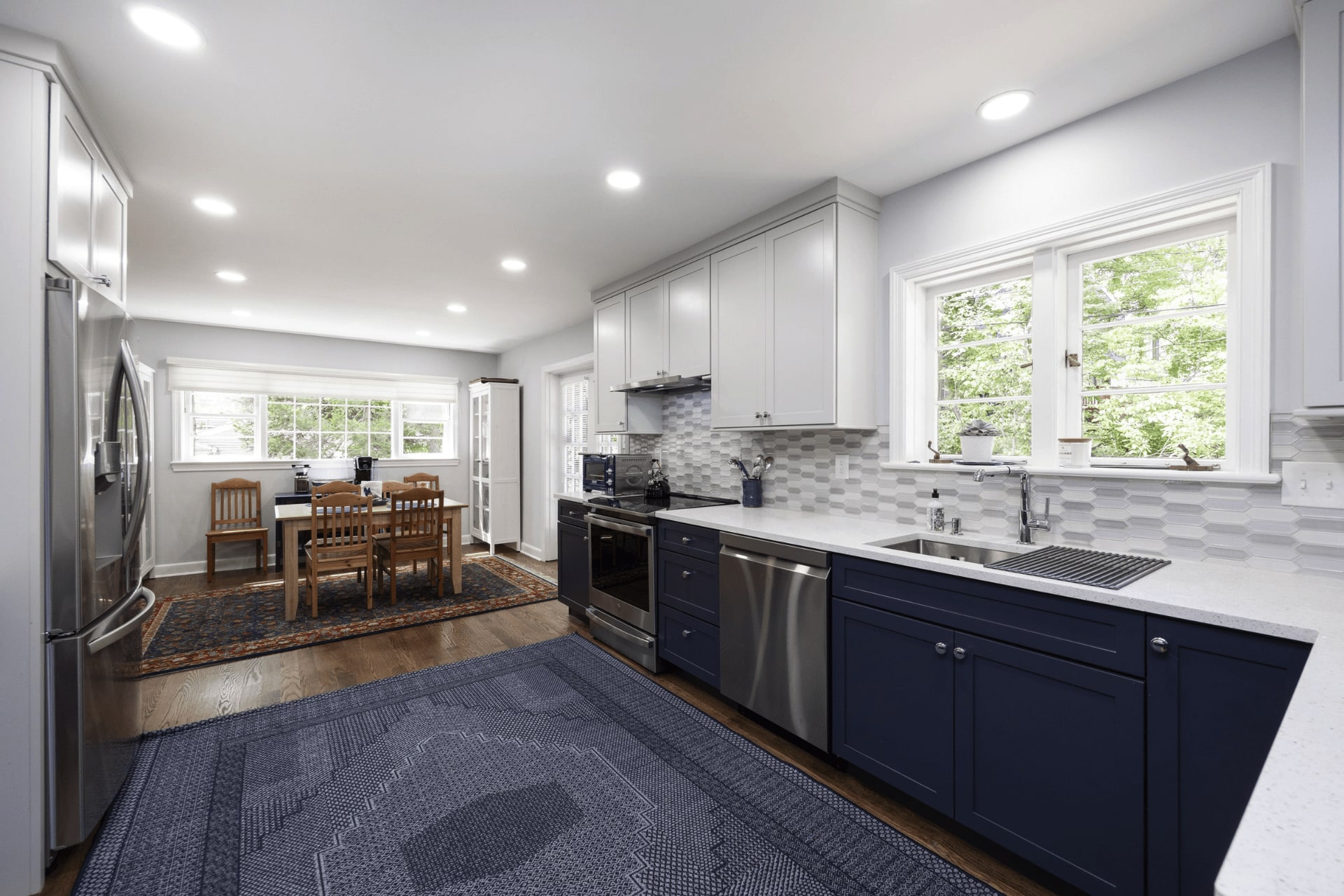 A modern kitchen features navy cabinets and stainless steel appliances. A countertop with a sink is central, while a dining area with wooden chairs and a patterned rug lies beyond. Natural light streams through large windows.