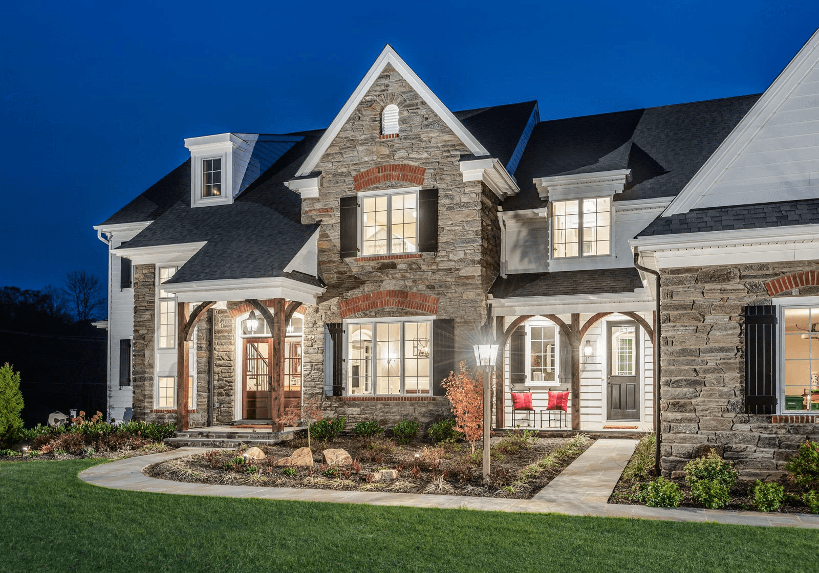 A large, illuminated stone house features an inviting entrance with wooden arches. The porch is decorated with red chairs, surrounded by landscaped greenery under a dark blue sky.