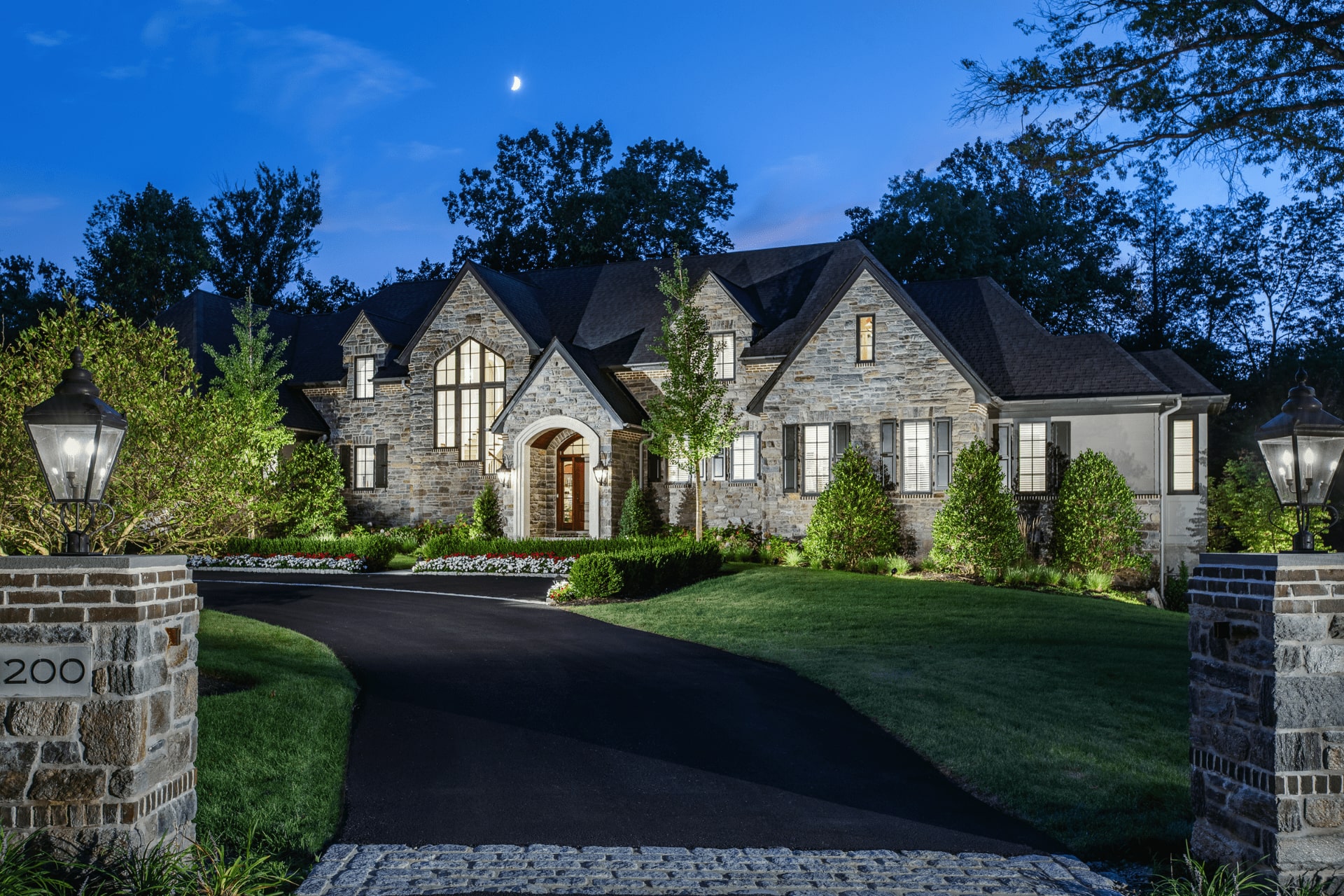 A stone house with a large arched entryway is illuminated against a twilight sky. Lush landscaping and a winding driveway lead to the residence, flanked by decorative lamps.