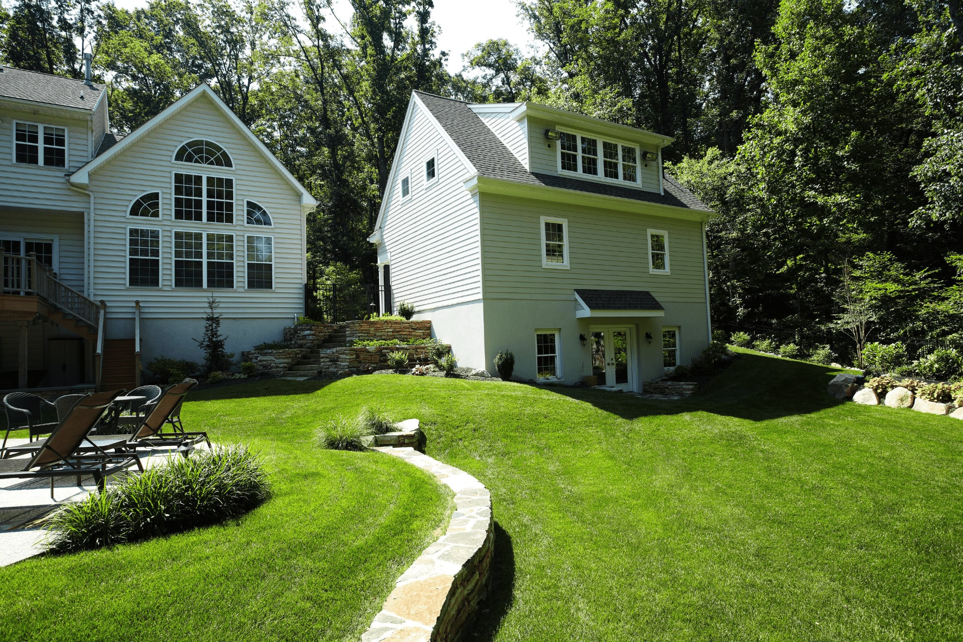 A two-story house with a light exterior is surrounded by a manicured lawn and trees. A stone pathway curves through the green grass, leading toward the house's entrance.