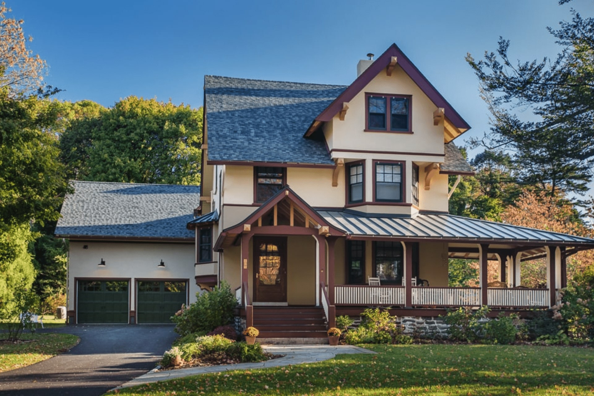 A charming, two-story house with a gabled roof and a porch features decorative wooden accents. It sits in a lush, green yard with trees and a paved driveway nearby.