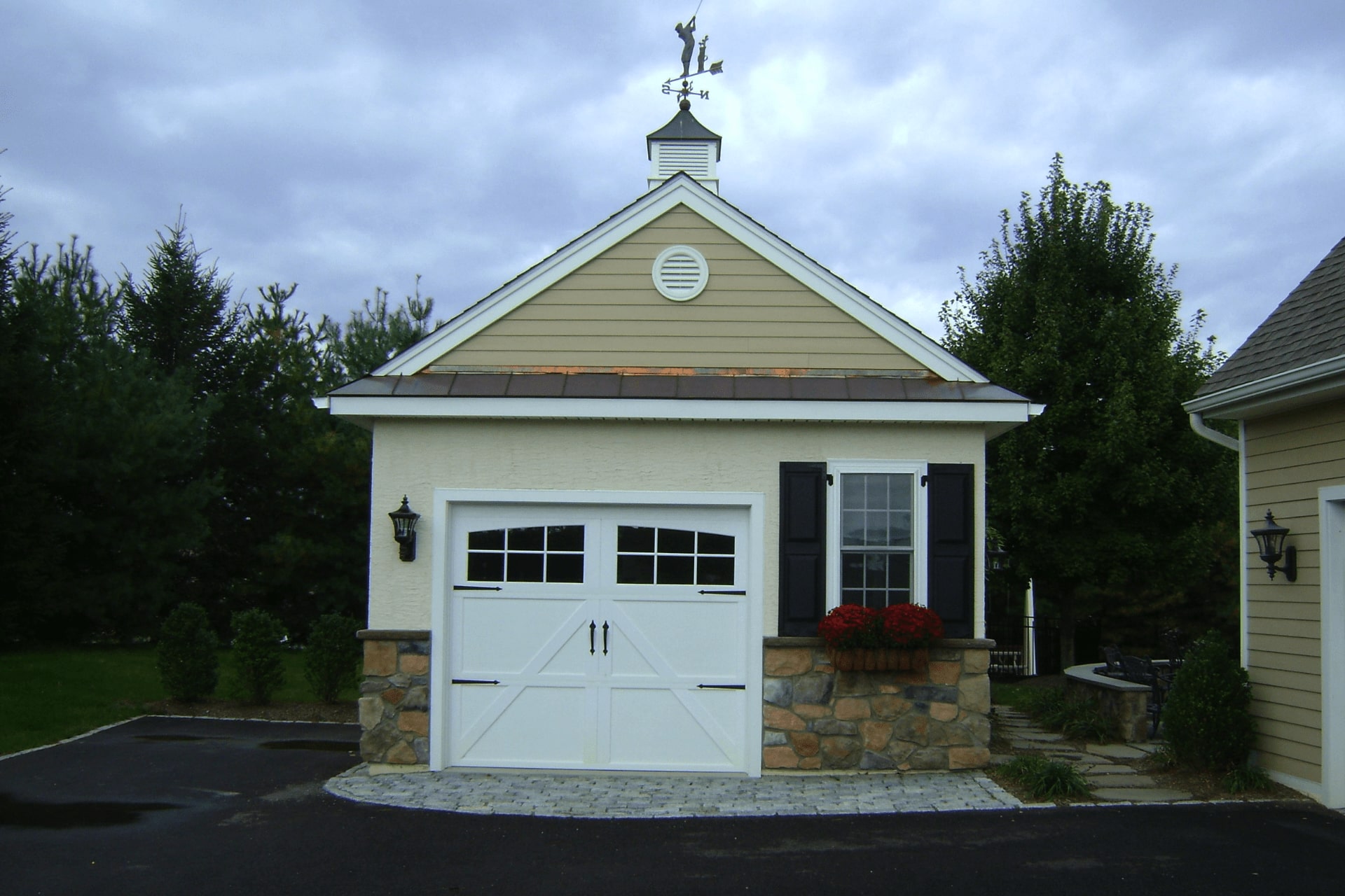 A small garage features a white, double door with windows, stone accents, and black shutters. It sits on a paved driveway surrounded by greenery under an overcast sky.