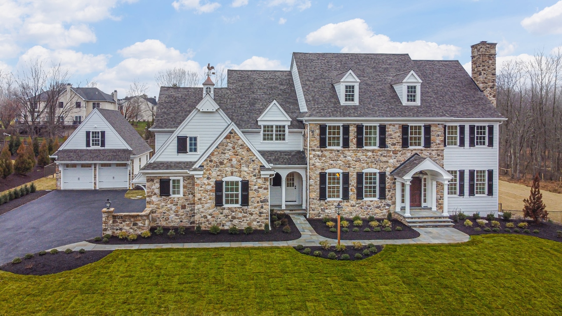 A large, two-story house features a mix of stone and siding, with multiple windows and a central entrance. It is set in a spacious yard, surrounded by trees and a paved driveway.