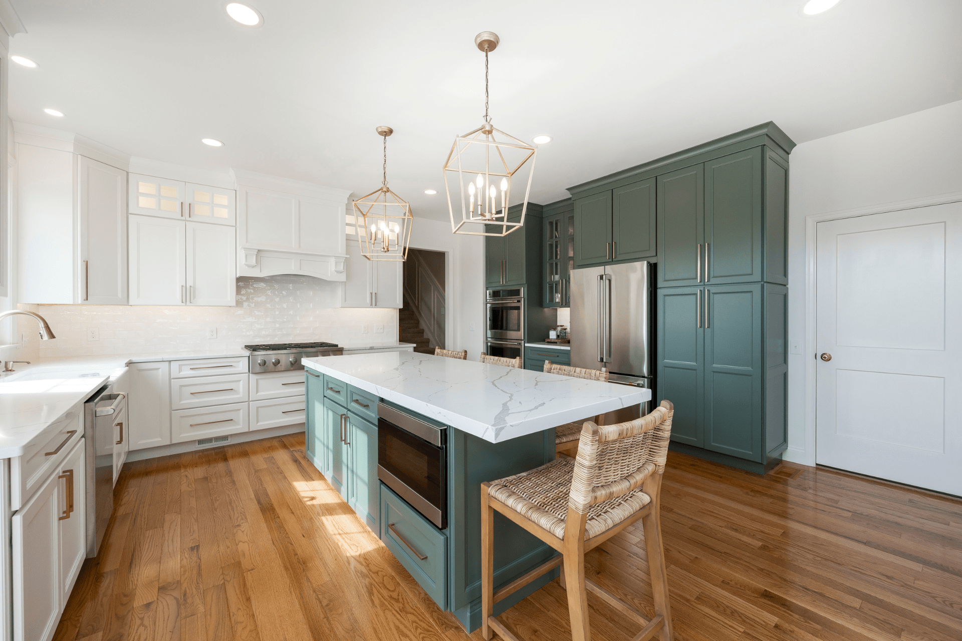 A modern kitchen features a central island with a marble countertop, a gas stove, and wooden flooring. Pendant lights hang above, complemented by white and dark green accent cabinetry.