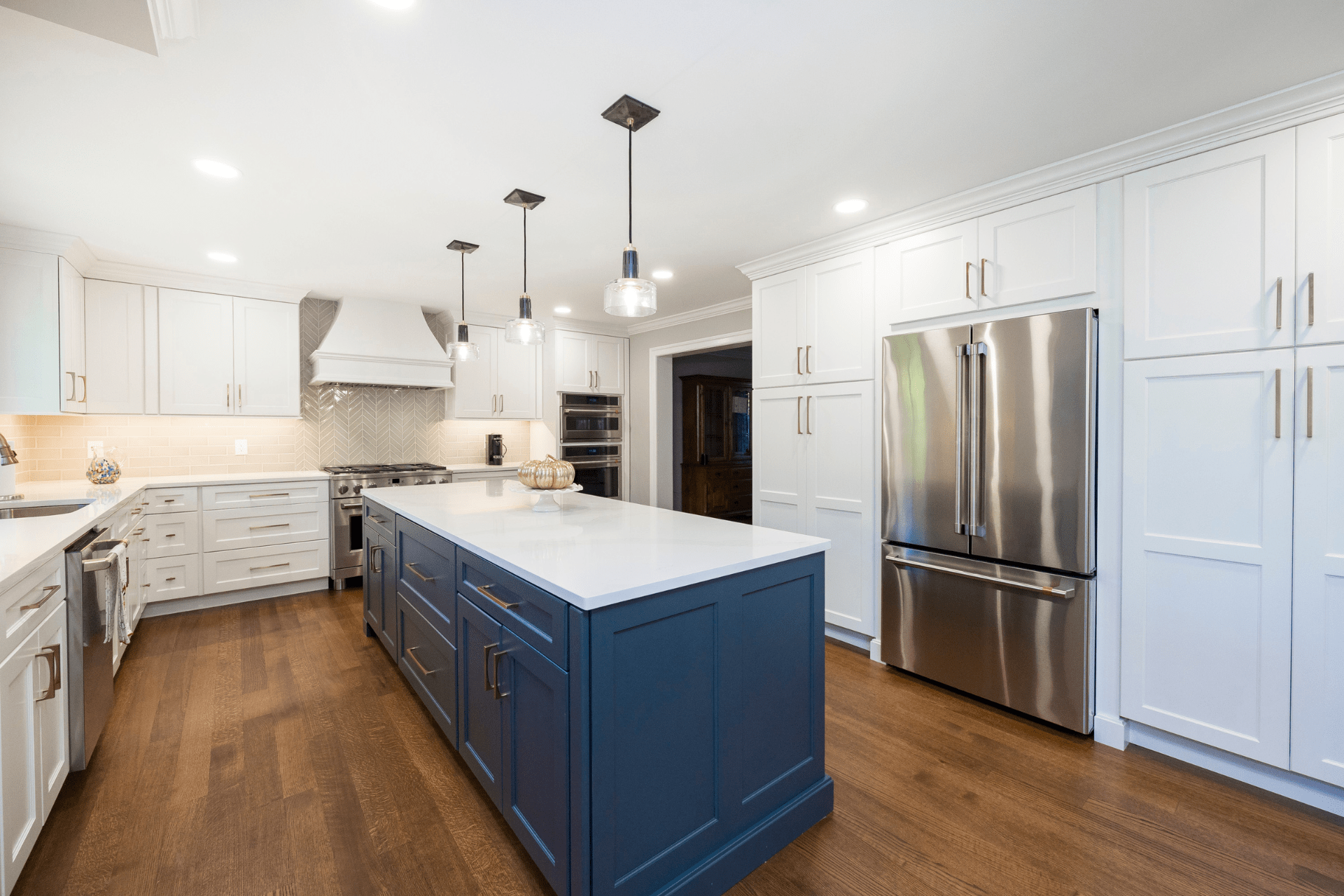 A modern kitchen features a large blue island with a white countertop. Two-tone pendant lights hang above. White cabinetry and stainless steel appliances create a sleek, bright atmosphere with hardwood flooring.