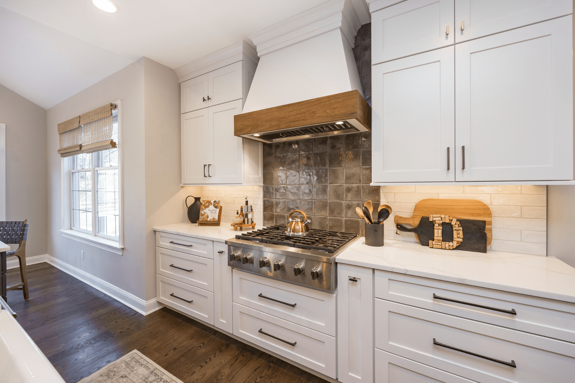 A modern kitchen features a gas stove with a metallic finish, a wooden range hood above, and white cabinets. Natural light enters through a window, casting a warm glow on the countertops.