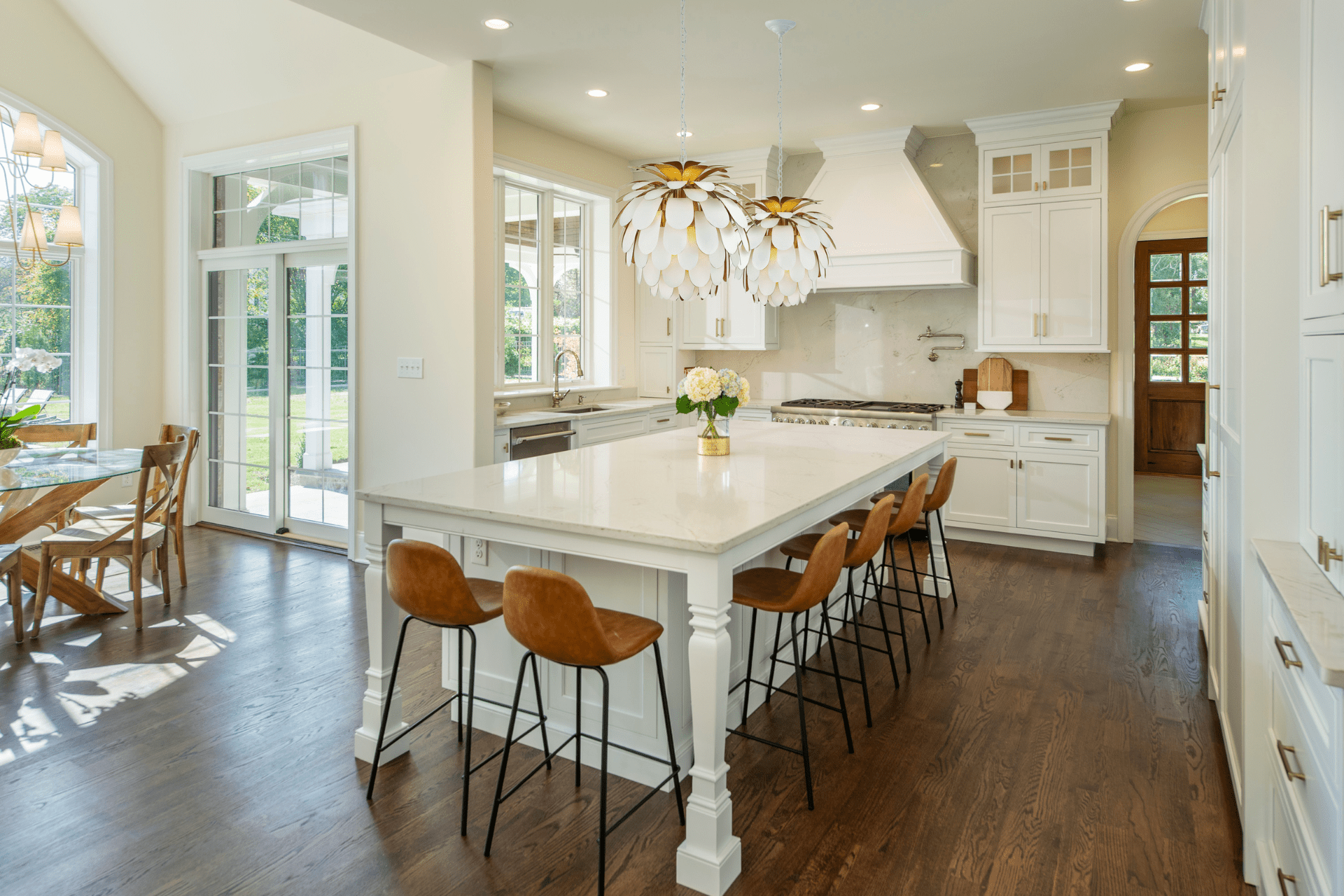 A modern kitchen features a large white island with leather stool seating. Elegant, petal-shaped light fixtures hang above, while a wood door and large windows brighten the space.