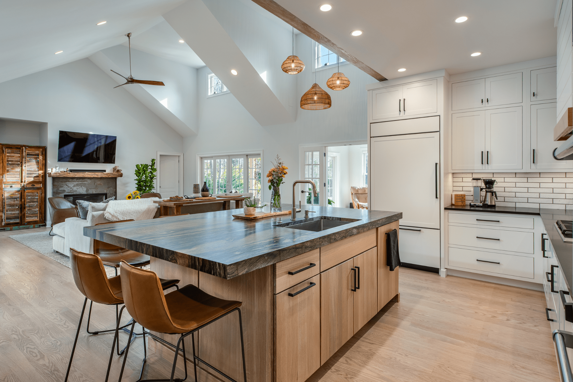 A wooden kitchen island with a sink sits prominently, while three brown chairs are placed around it. Modern appliances and bright lighting complement a spacious, airy dining area in a home setting.