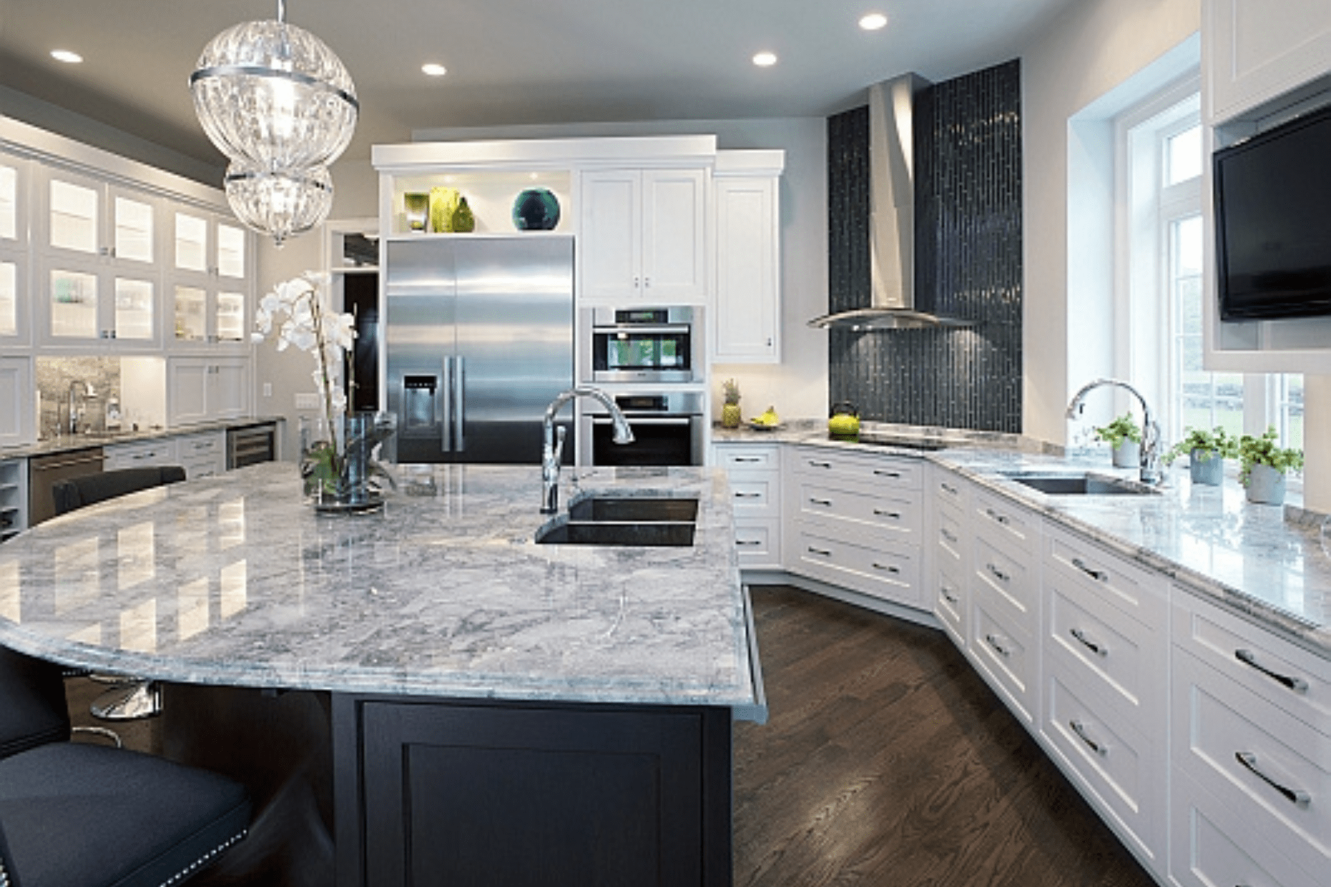 A modern kitchen features a large, curved granite countertop with a sink, surrounded by white cabinetry and clean stainless steel appliances. Decorative plants enhance the sleek, elegant atmosphere.