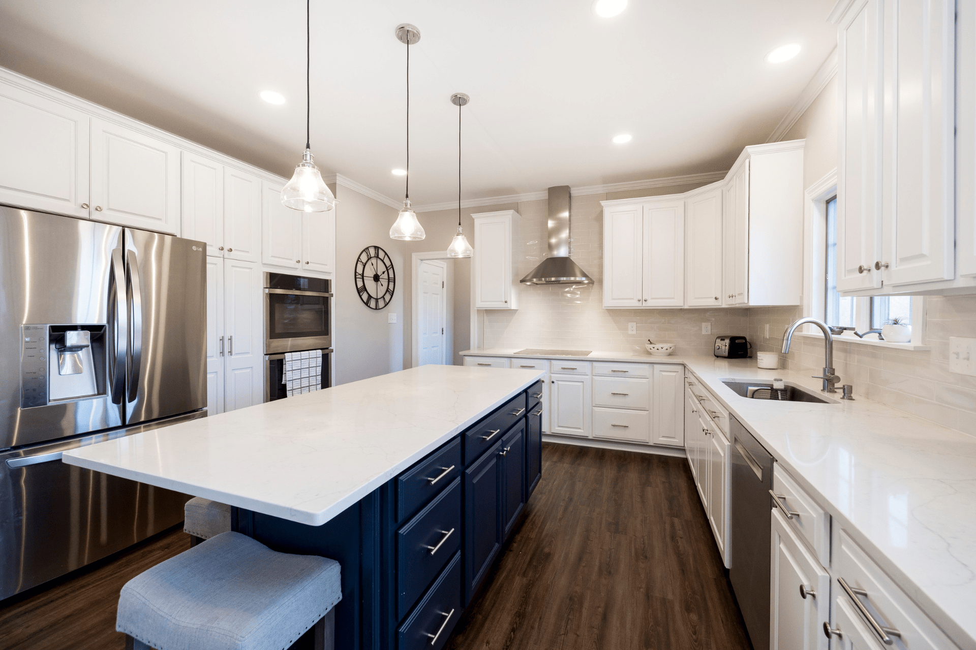 A modern kitchen features a large marble island with navy cabinets, stainless steel appliances, and white cabinetry, illuminated by minimal pendant lights, with warm wood flooring and a clock on the wall.