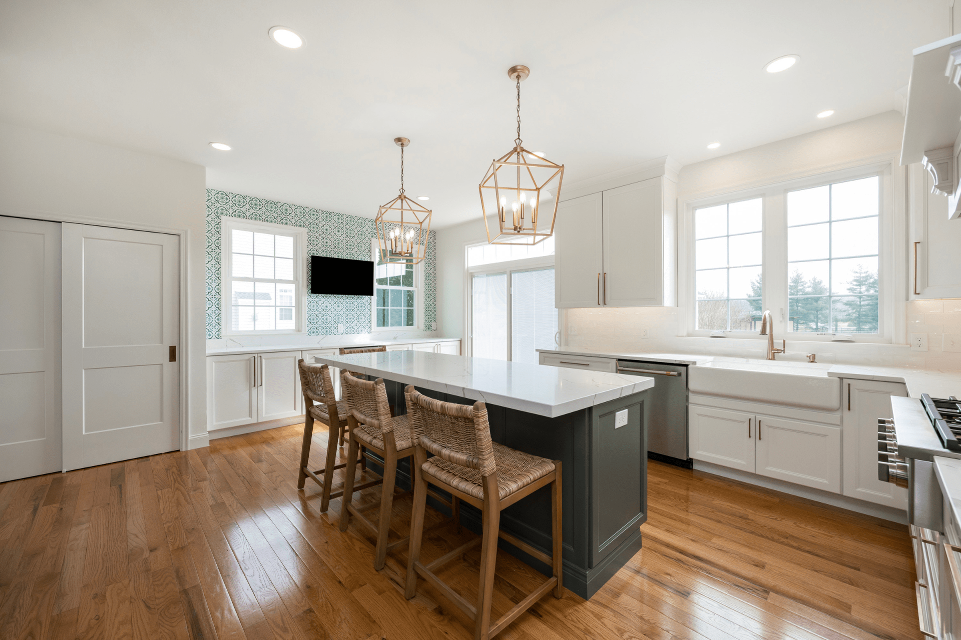 A modern kitchen features a long island with beige wicker stools. It has light-toned cabinets, a large window, and decorative green wallpaper. Two geometric pendant lights hang above, illuminating the space.