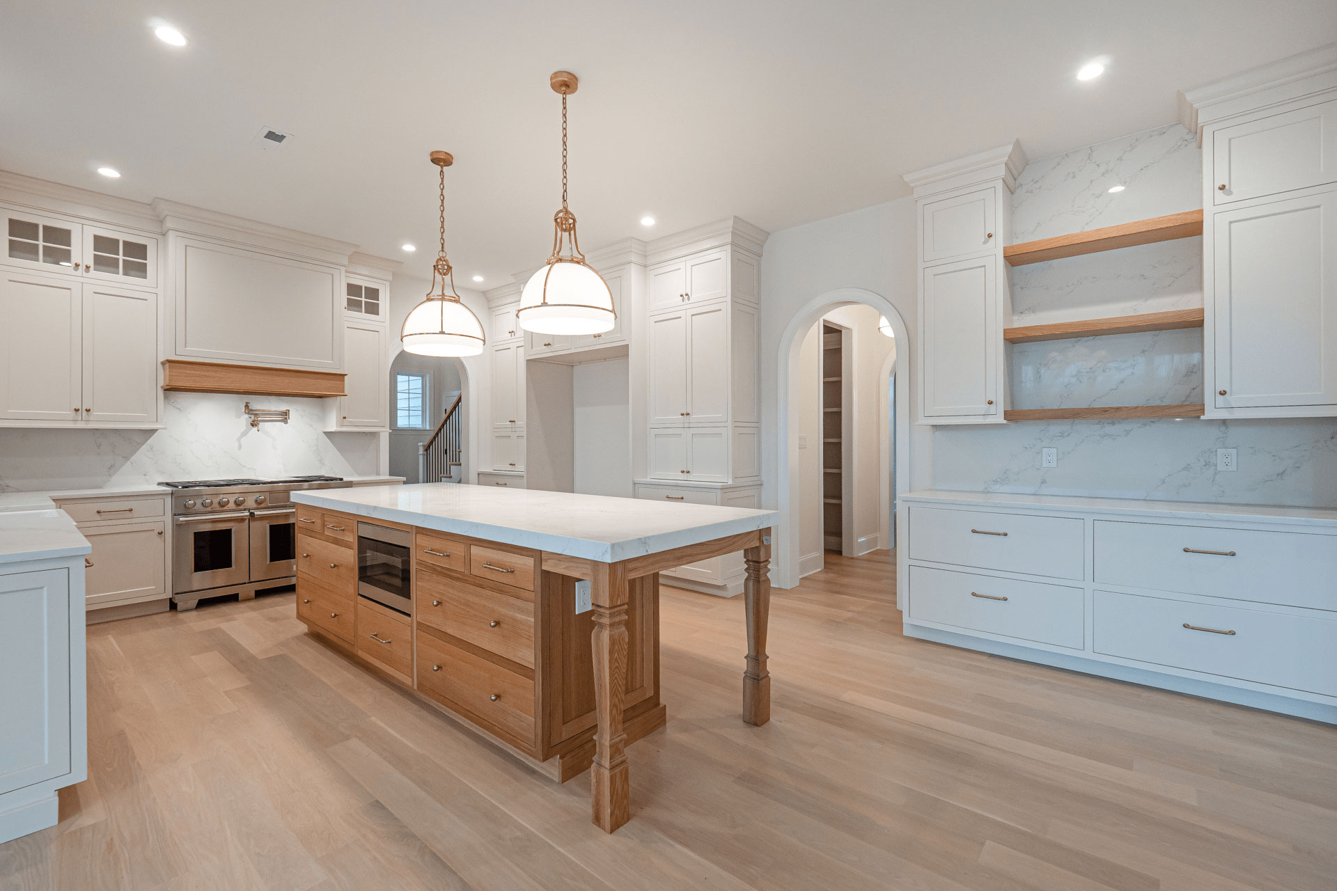 Spacious modern kitchen with white cabinetry, marble countertops, light wood floors, a wooden island with built-in oven, two pendant lights, open shelves, and an arched doorway.