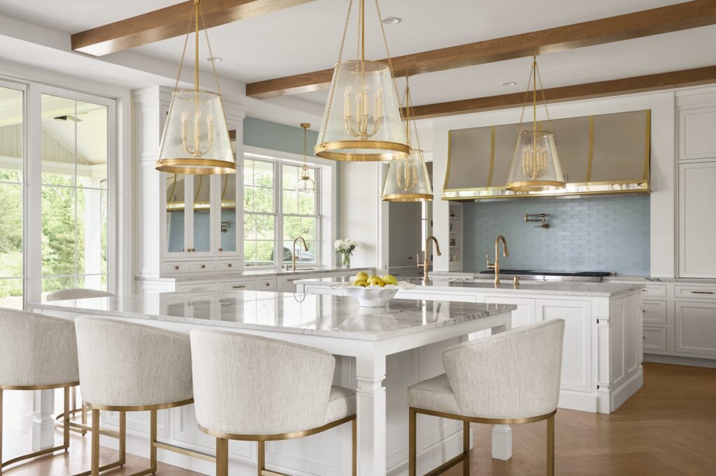 Modern kitchen with white cabinets, marble countertops, gold accents, and three large glass pendant lights over an island with beige upholstered barstools. Large windows let in natural light, and a bowl of lemons sits on the island.