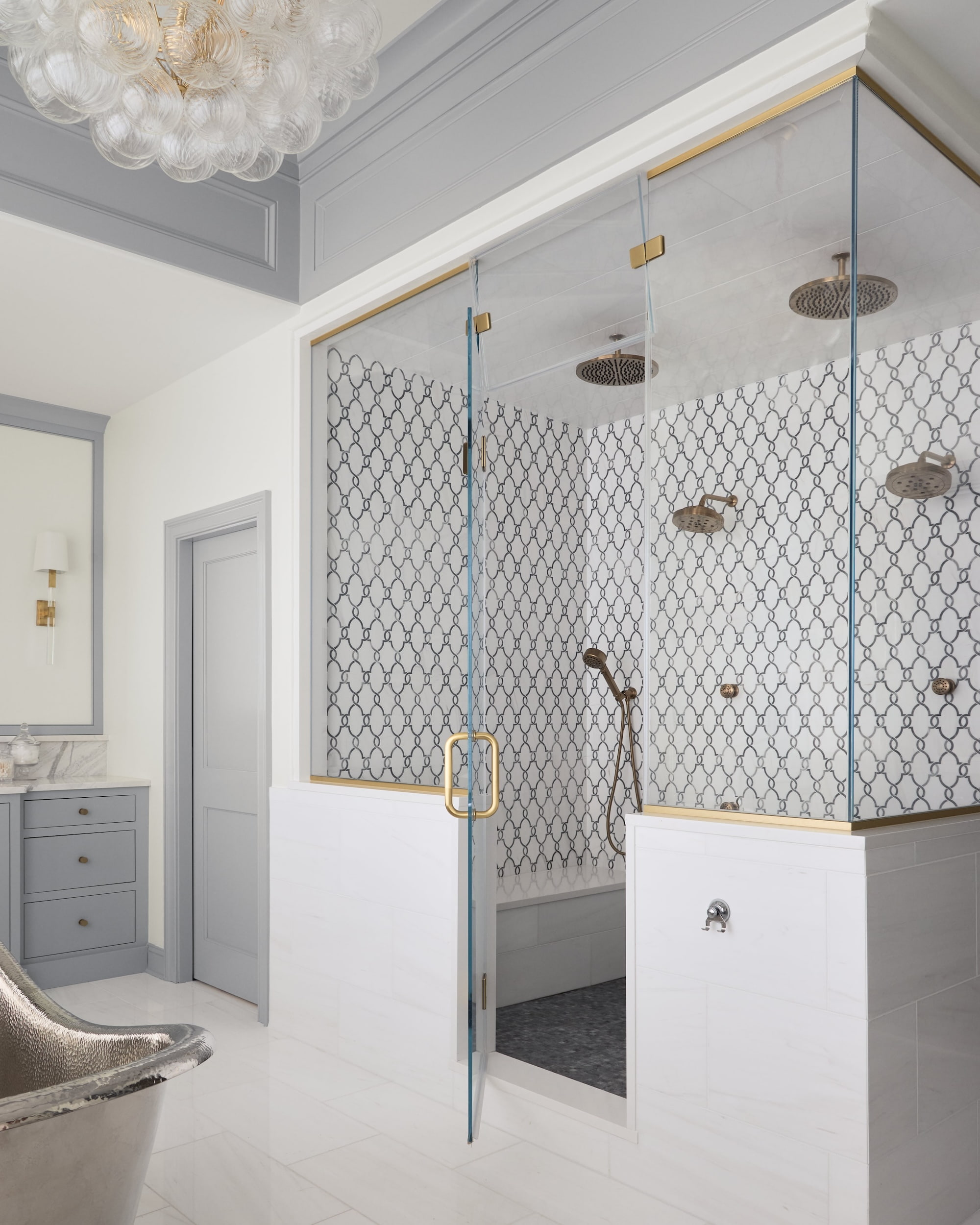 A modern bathroom with a glass-enclosed shower featuring geometric patterned tiles, gold fixtures, and a double showerhead; grey cabinets, a metallic freestanding tub, and a chandelier complete the elegant space.