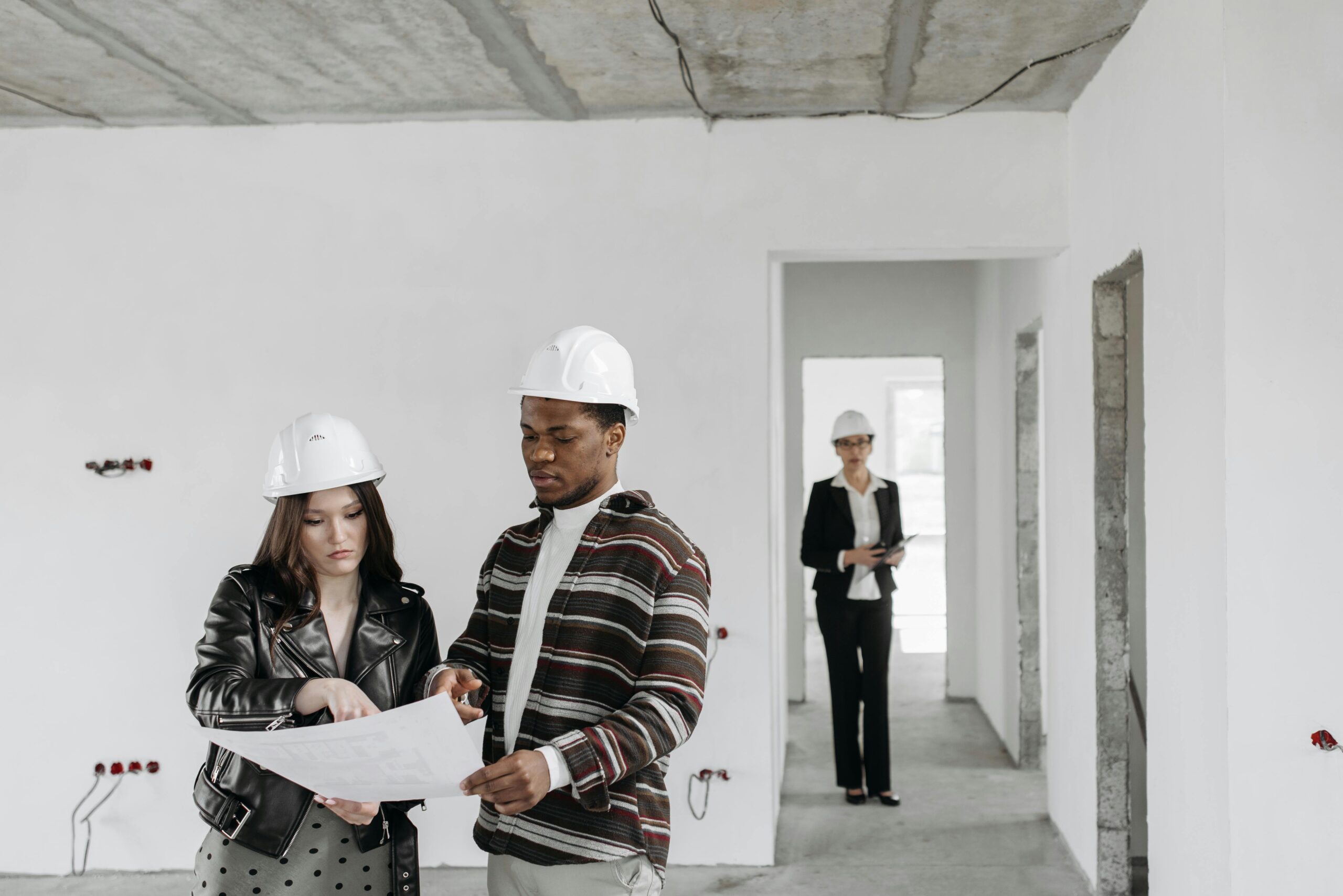Two people wearing hard hats review building plans in an unfinished room, while a third person in a suit and hard hat stands in the background holding documents. The space has exposed concrete ceilings and white walls.
