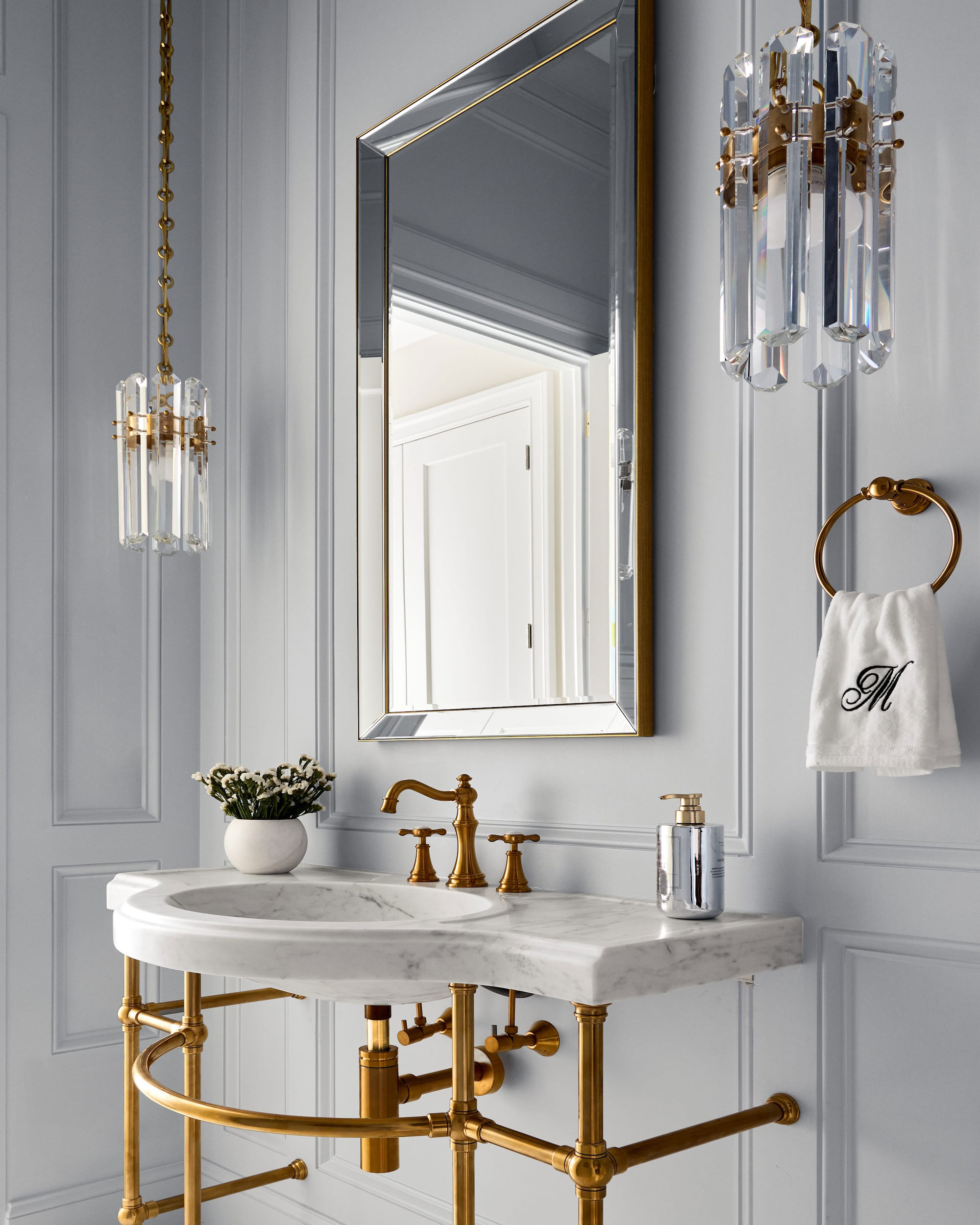 Elegant bathroom with gold fixtures, a marble sink, a wall-mounted mirror, glass pendant lights, and a white hand towel with a monogrammed M on a gold ring. A small plant sits on the counter.