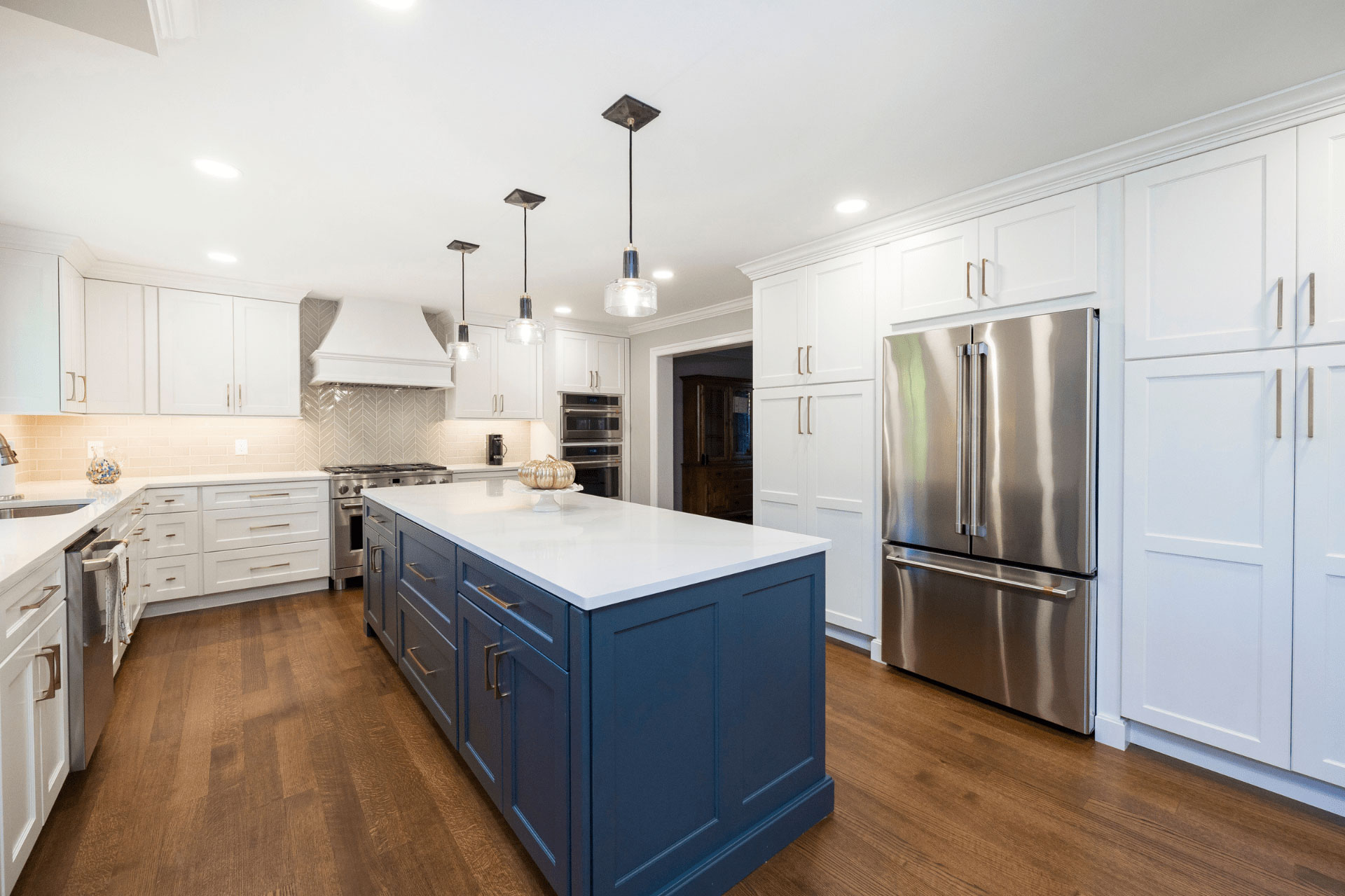 Modern kitchen with white cabinets, stainless steel appliances, a large blue island with a white countertop, pendant lights, and hardwood flooring.