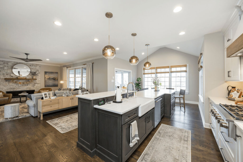 Modern open-concept kitchen and living area with a large island, pendant lights, dark wood floors, light cabinetry, and large windows letting in natural light. The living room has a sofa, chairs, and a fireplace.