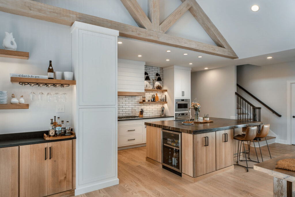 Modern kitchen with light wood cabinets, a black countertop island with bar stools, open shelves with dishes and glasses, and a built-in oven. Exposed wooden beams and white brick backsplash add character.