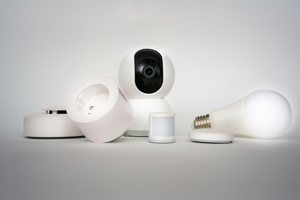 A group of smart home devices, including a security camera, light bulb, motion sensor, plug, and other wireless devices, arranged on a plain white surface with a neutral background.
