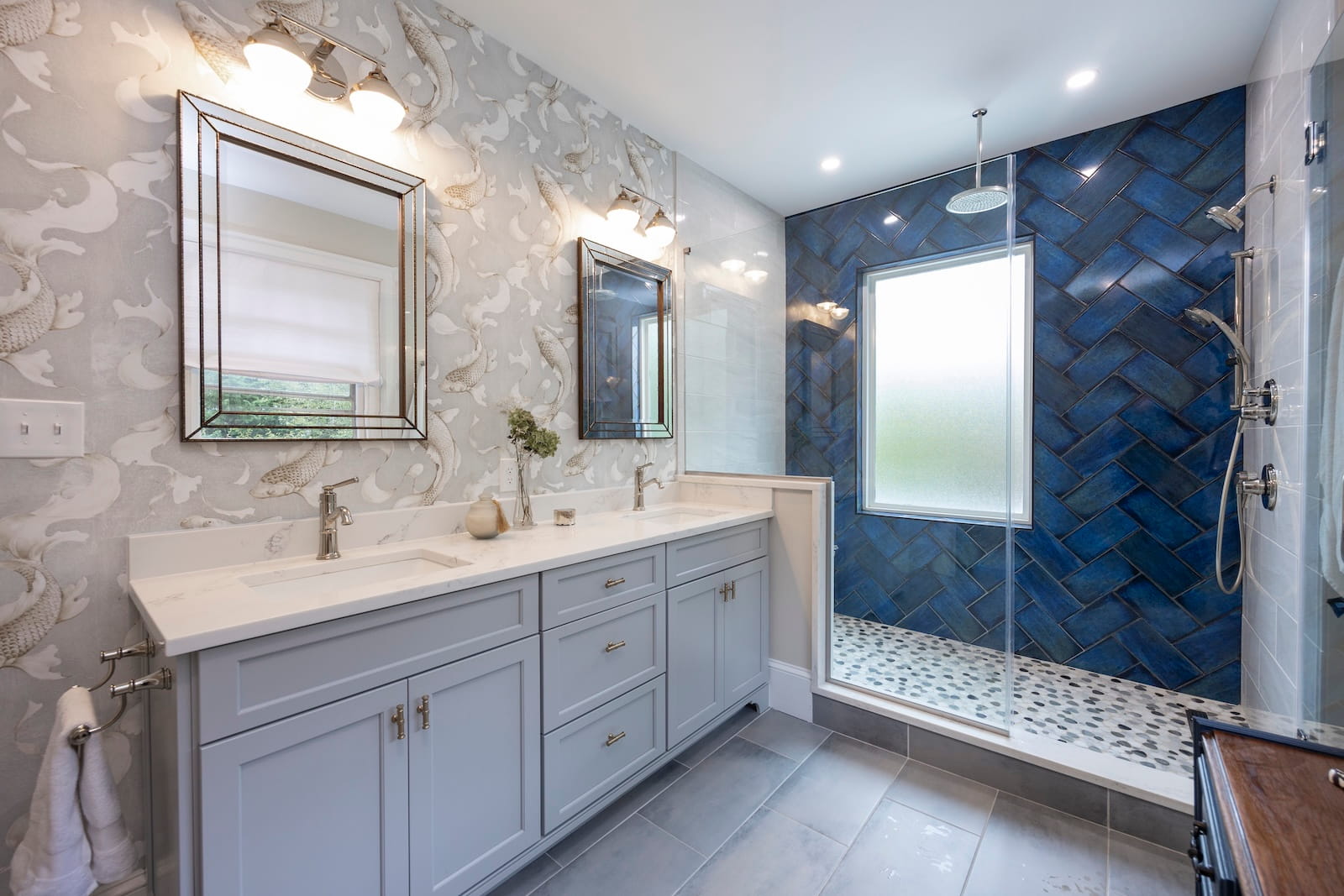 Modern bathroom with light gray double vanity, two mirrors, decorative wallpaper, and a glass-enclosed shower featuring a blue herringbone tile wall and pebble tile floor, with natural light from a frosted window.