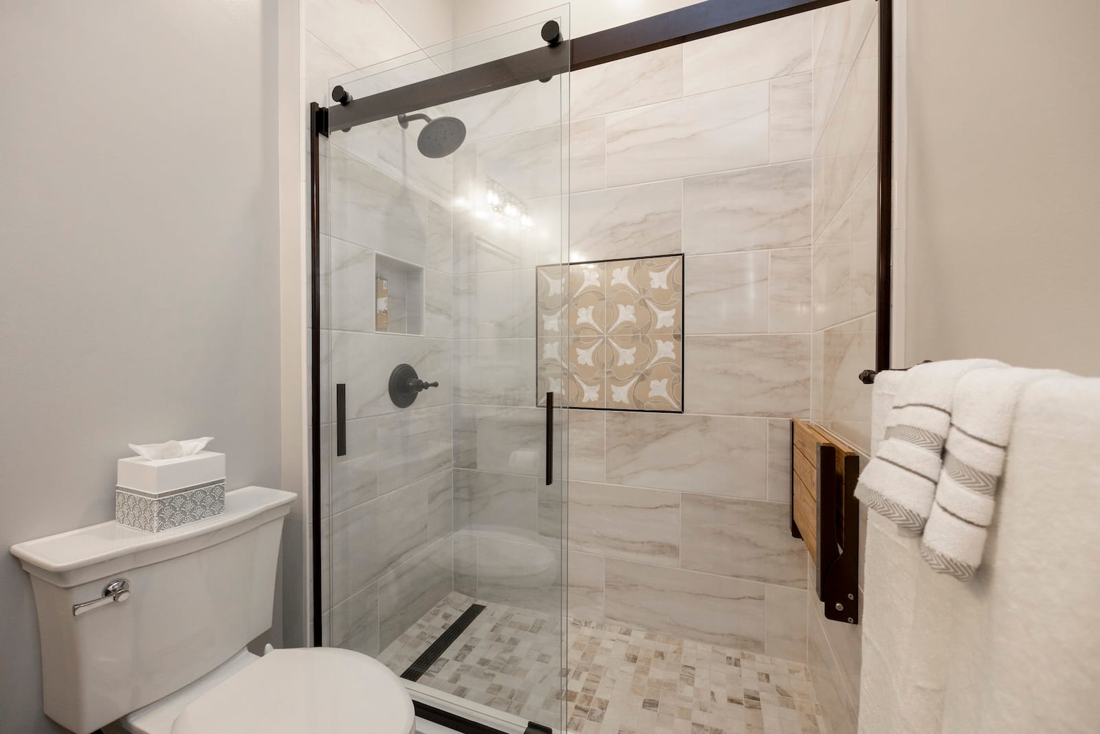 Modern bathroom with a glass-enclosed shower featuring beige tiles, a decorative accent tile pattern, a rain showerhead, and built-in shelf. A toilet and folded white towels are visible in the foreground.