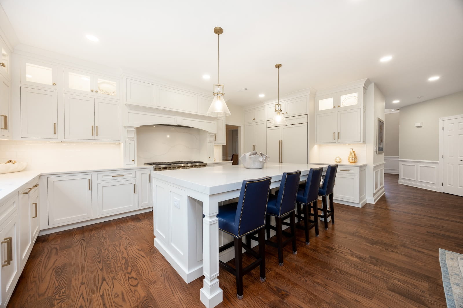 Bright, spacious kitchen with white cabinets, a large island with a white countertop, four navy blue chairs, pendant lighting, hardwood floors, and built-in appliances.