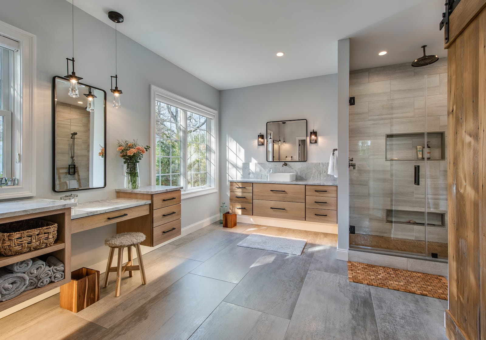 Modern bathroom with wood accents, large windows, double vanity, glass-enclosed shower, wall mirrors, pendant lights, baskets with towels, flowers in a vase, and natural light streaming in.