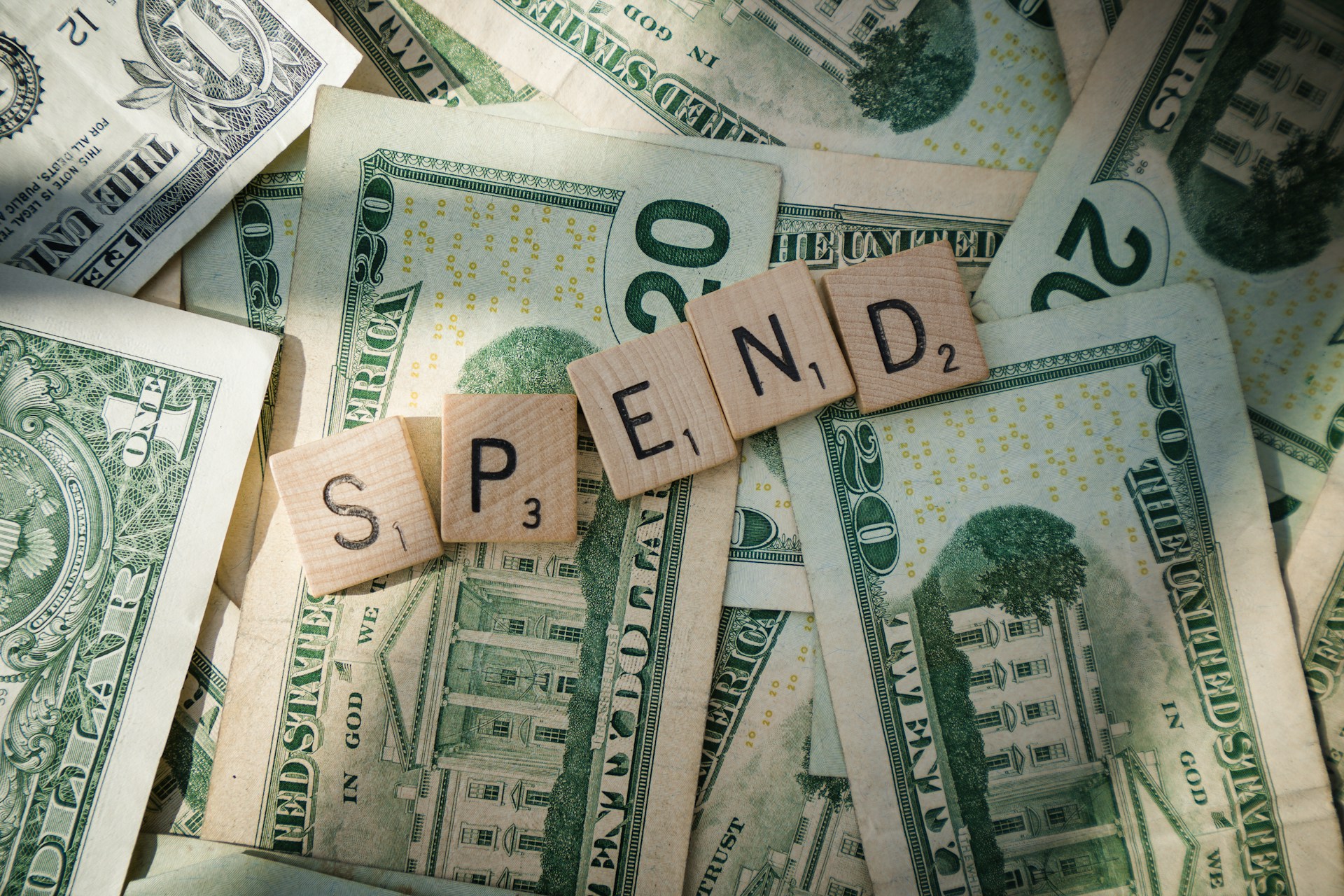 Scrabble tiles spelling SPEND are arranged on top of scattered US dollar bills, including $1, $10, $20, and $50 denominations, emphasizing the concept of spending money.