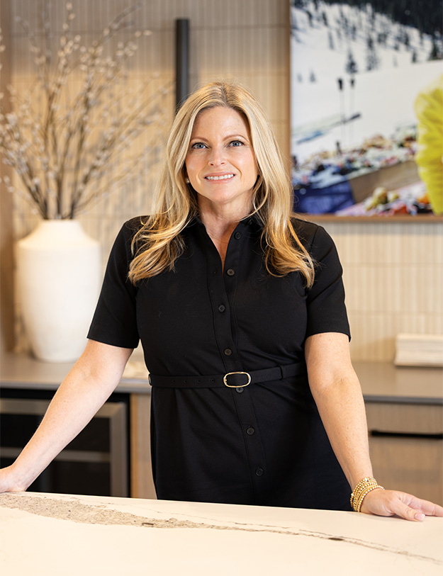 A woman with long blonde hair, wearing a black dress and gold bracelets, smiles while standing behind a marble counter in a modern room with a vase and a large photo on the wall.