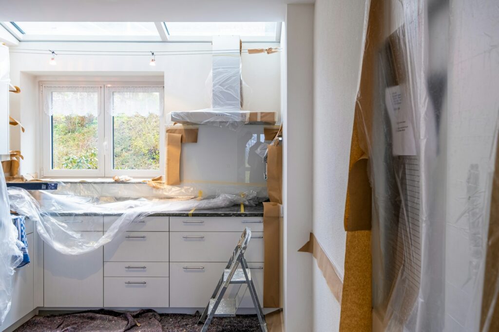 A kitchen under renovation, with cabinets, counters, and appliances covered in protective plastic. A ladder stands in the middle, and sunlight comes in through a skylight and window.