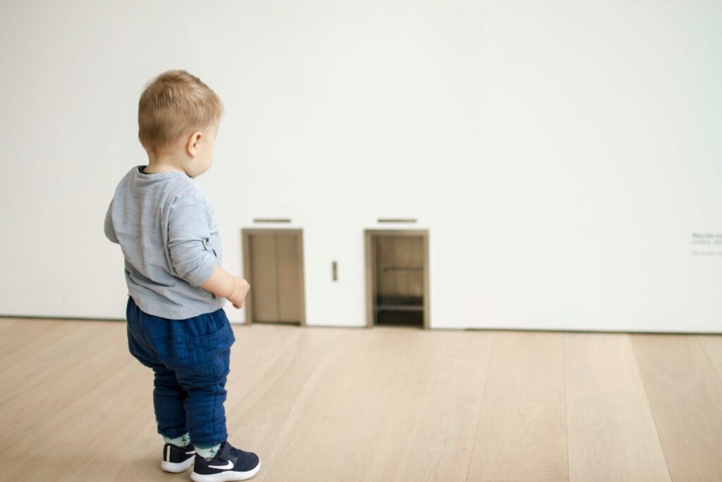 A toddler in blue pants and a grey shirt stands on a wooden floor, looking curiously at two small doors set into a plain white wall.